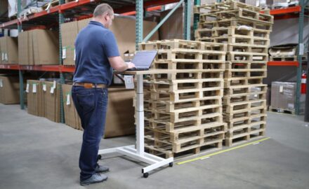 Photo of a man working on a laptop at a standing desk, in the aisle of a warehouse, next to two stacks of empty pallets.